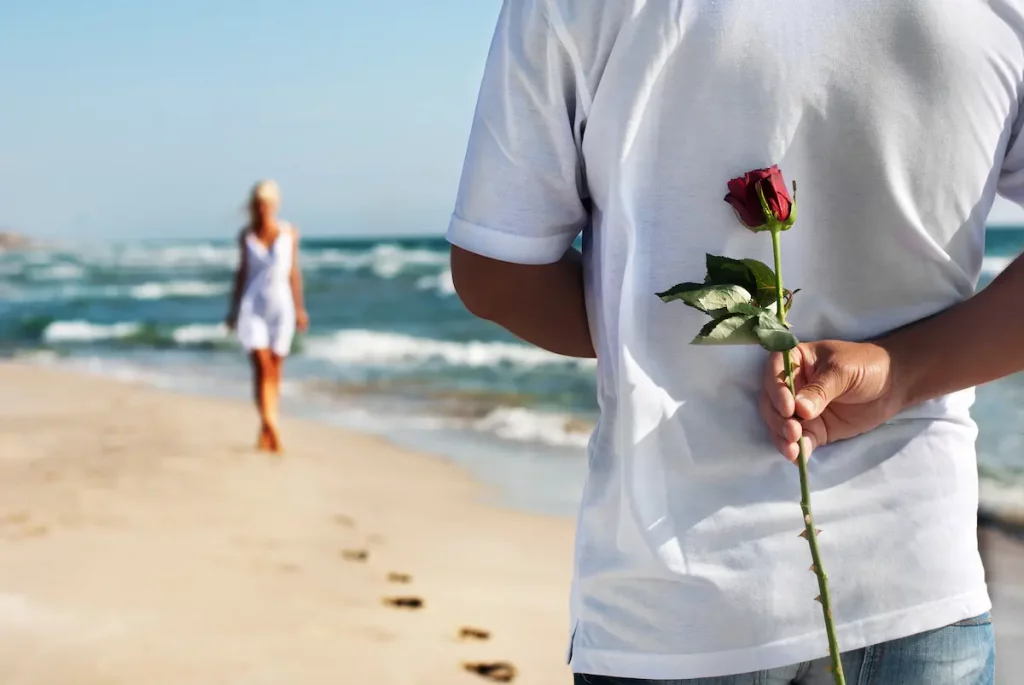 Younger man who is dating an older woman is holding a rose behind his back on the beach waiting for his date