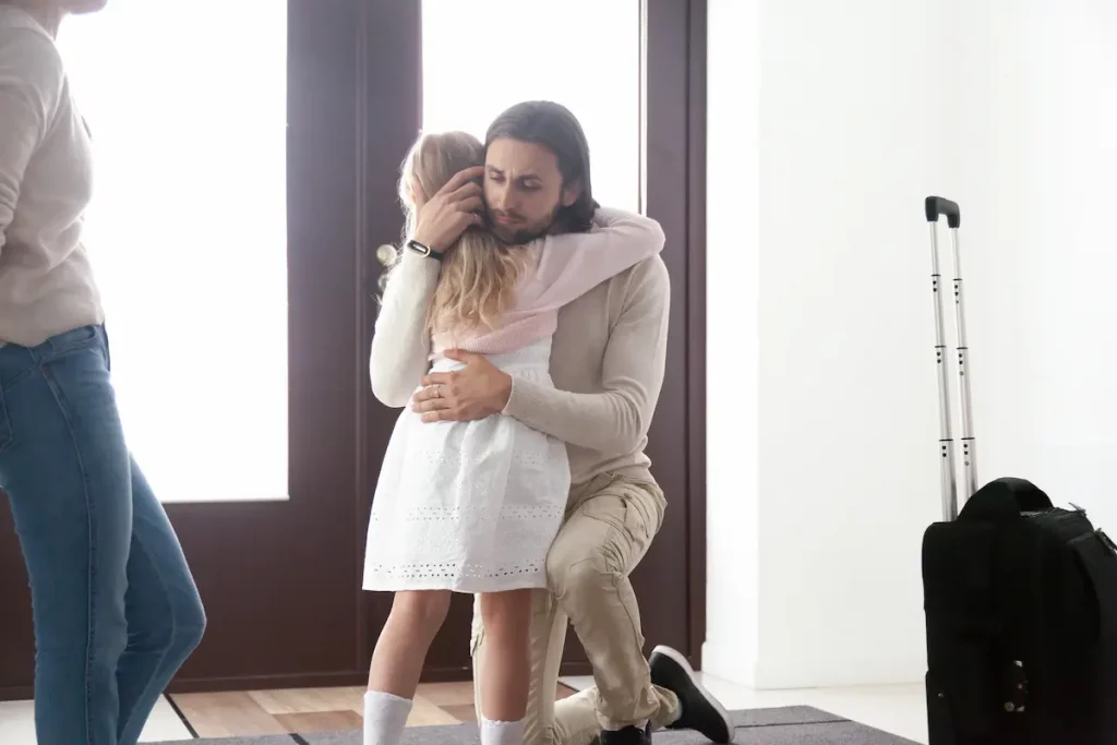 Absent father hugging his daughter before leaving house with luggage and mother stands off in distance watching