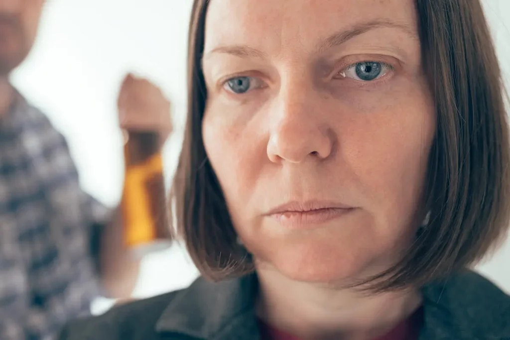 Close up of wife thinking how Husband Embarrasses Me with blurry man in background drinking from bottle of beer