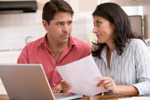 Husband and wife sitting at kitchen table with paper work and laptop looking at each other worried