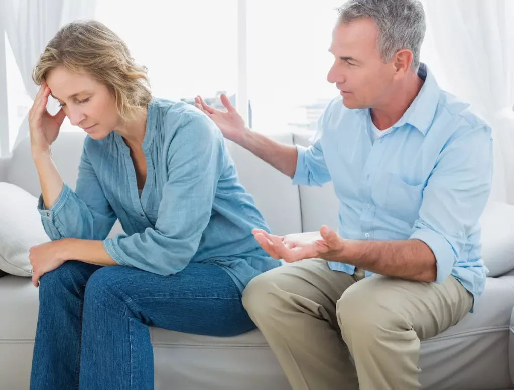 Angry wife refusing to talk to husband's mother sitting on couch with husband trying to compromise