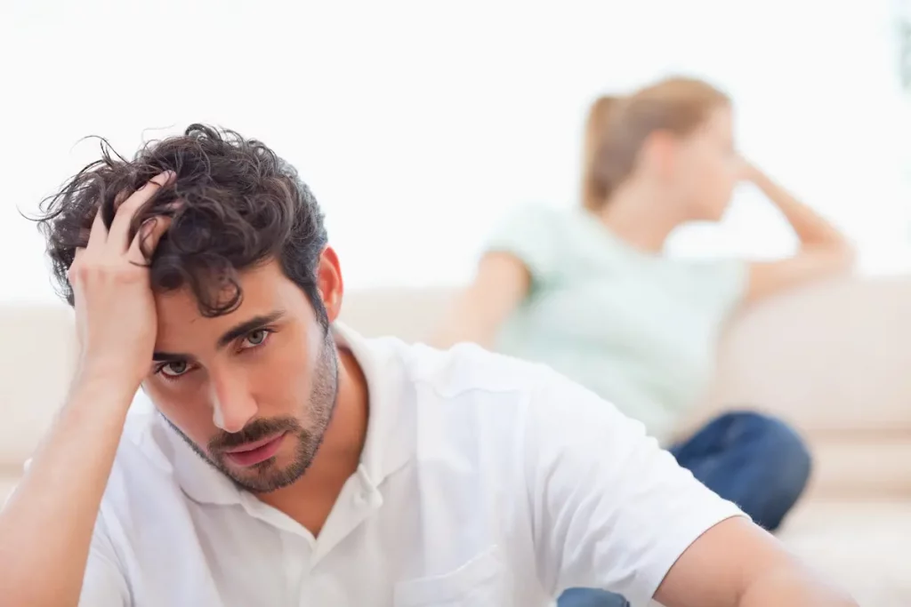 Upset husband with hand on forehead and his wife in background who never apologizes