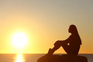 Woman sitting on rock in water looking at sunset reflection on when her family hurt her
