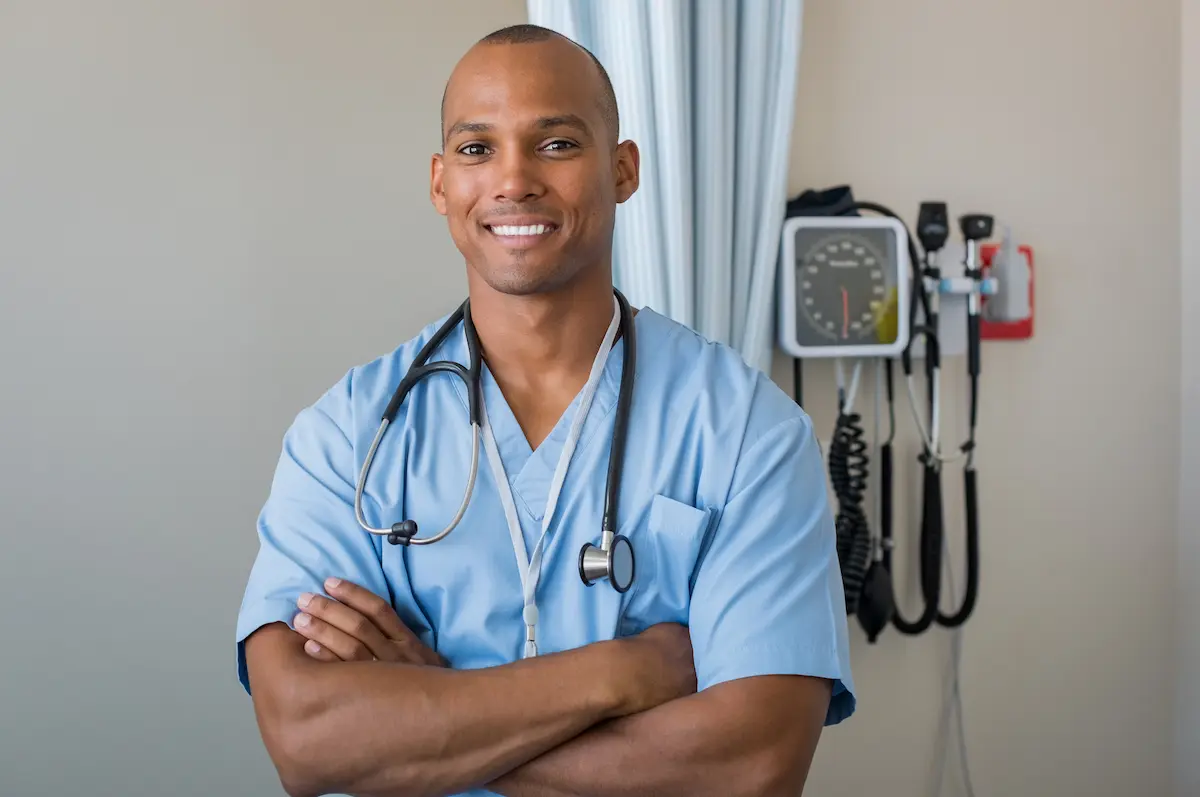 Male nurse who recently graduated nursing school posing in front of medical equipment with hands crossed on chest looking proud