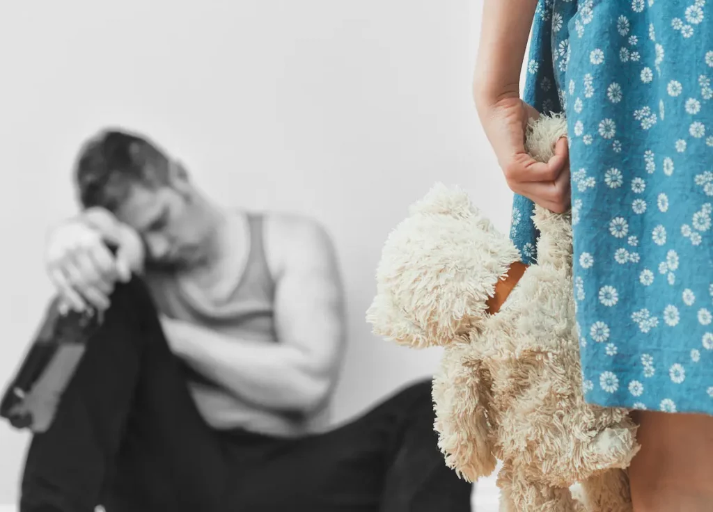 Daughter holding teddy bear while looking at her drunk deadbeat dad passed out on floor