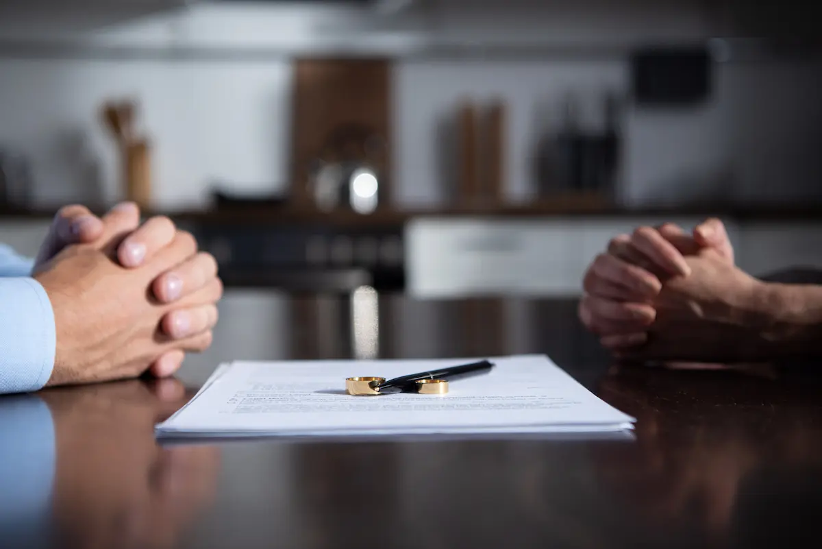 Couple sitting across table with hands crossed and wedding rings off on divorce work paper