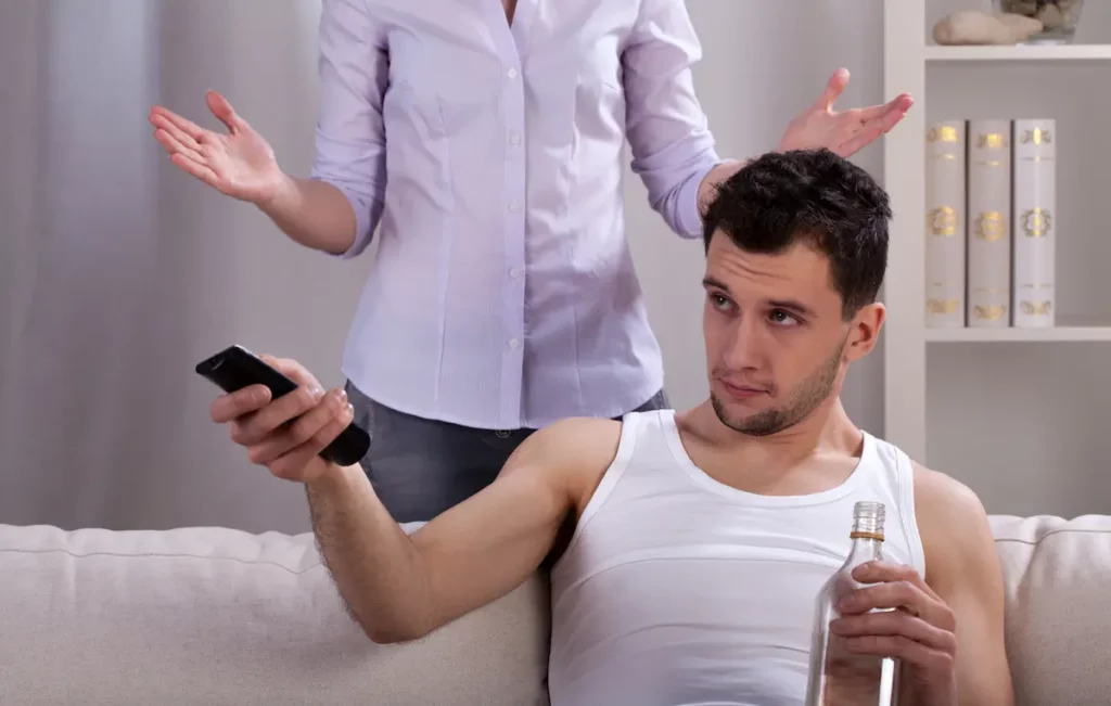 Lazy father sitting on couch with wife looking frustrated behind him