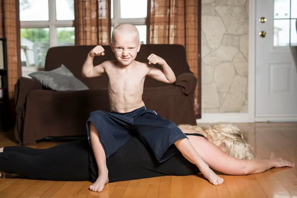 permissive parent exhausted on ground while child sits on top flexing muscles